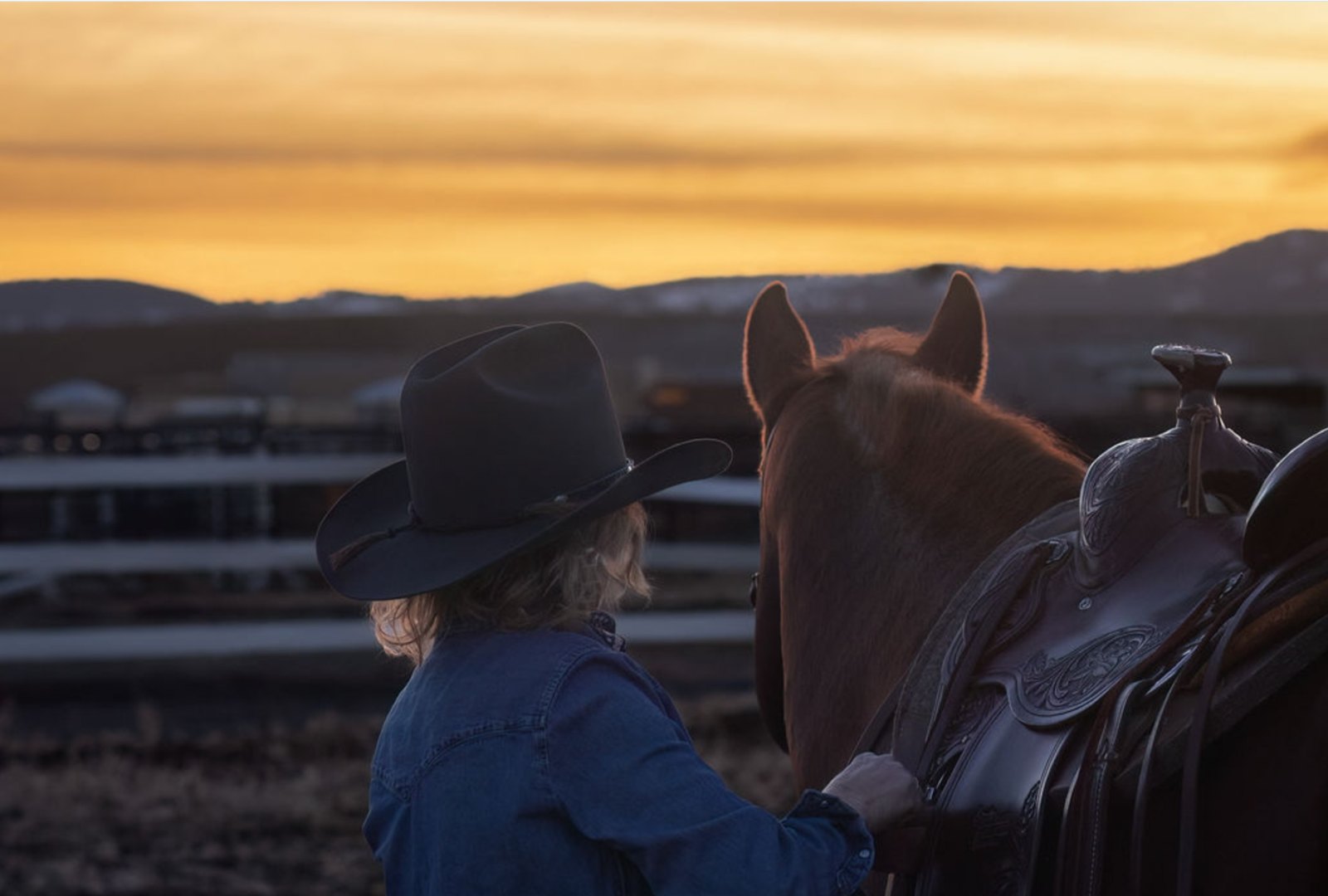 Cowgirl standing beside a saddled horse at sunset
